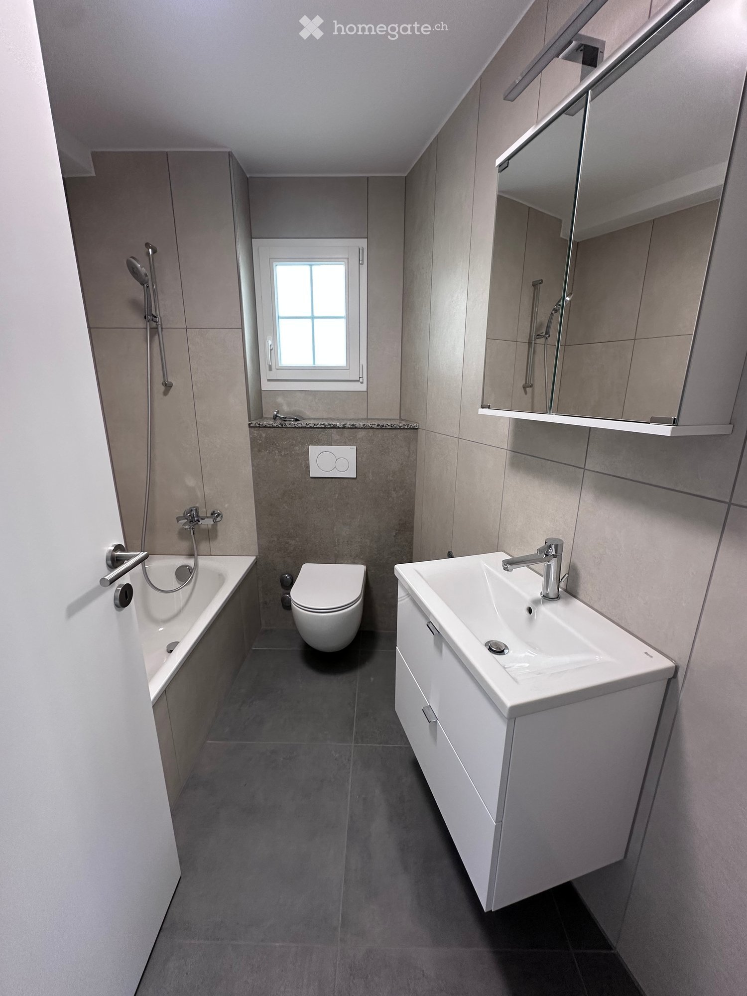 Modern bathroom with a bathtub, toilet, sink, and mirror. Features grey tiles and a window for natural light.