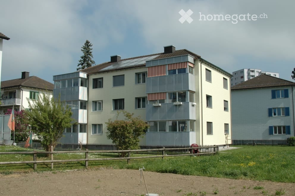 Multi-story residential building with balconies, solar panels on the roof, and surrounding greenery.
