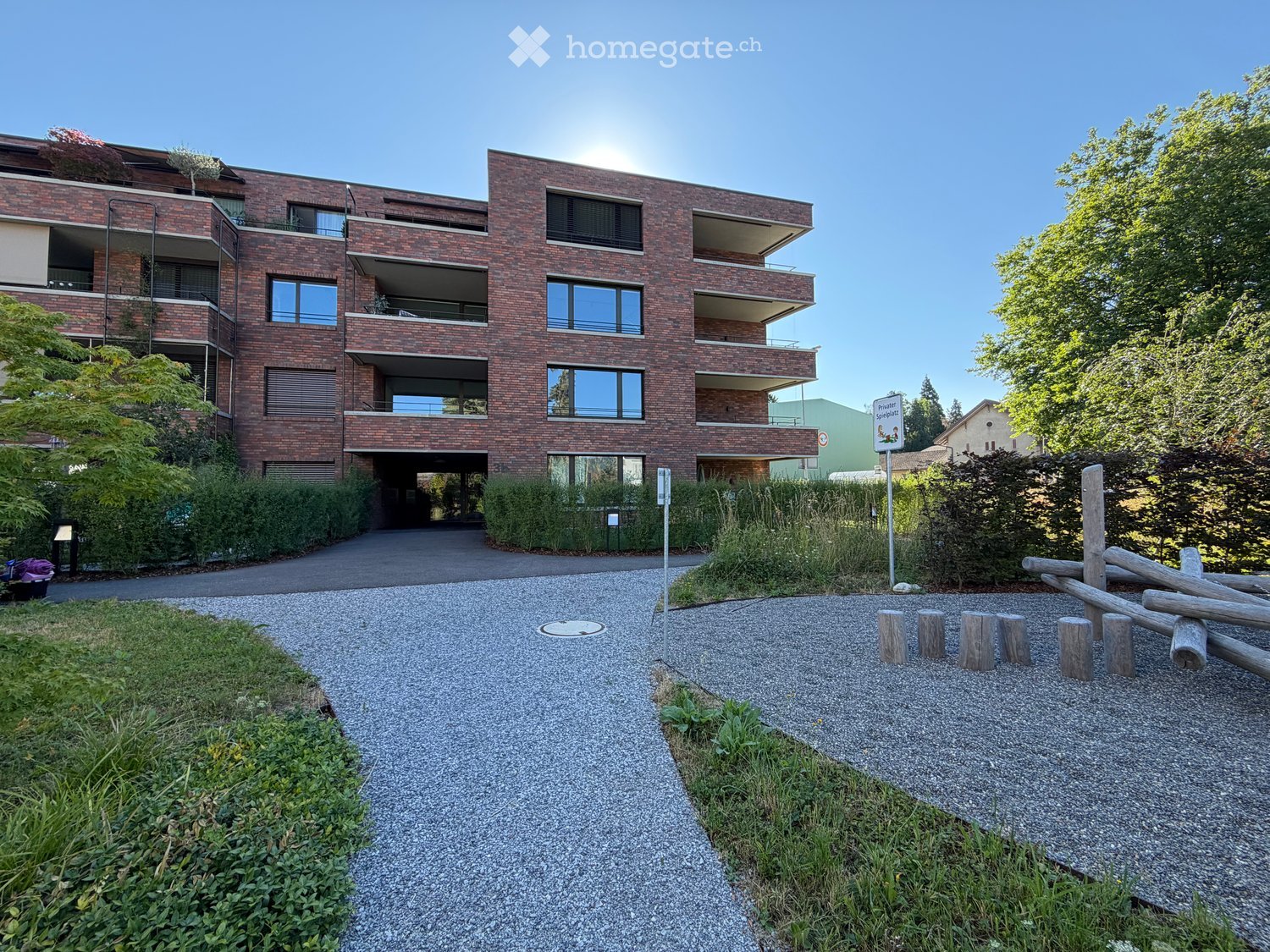 Modern multi-story brick building with balconies and large windows, surrounded by greenery and a gravel pathway.