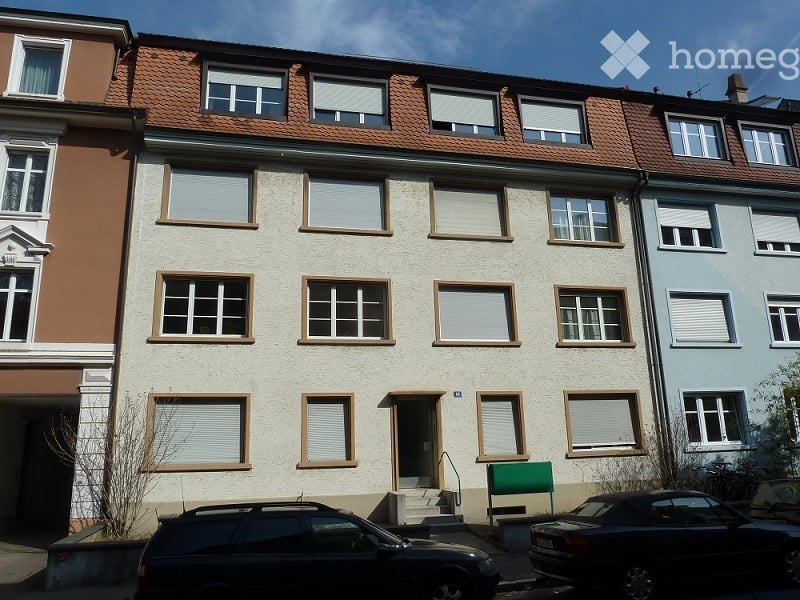 Multi-story residential building with beige facade, multiple windows with shutters, and a small entrance staircase leading to the main door.