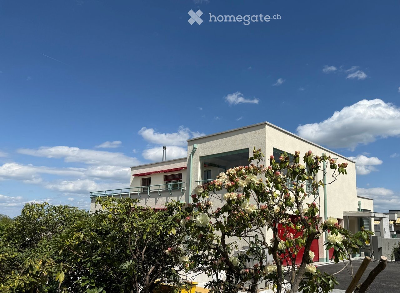 Modern single-story building with white walls, large windows, and a balcony overlooking a garden area with flowering shrubs.