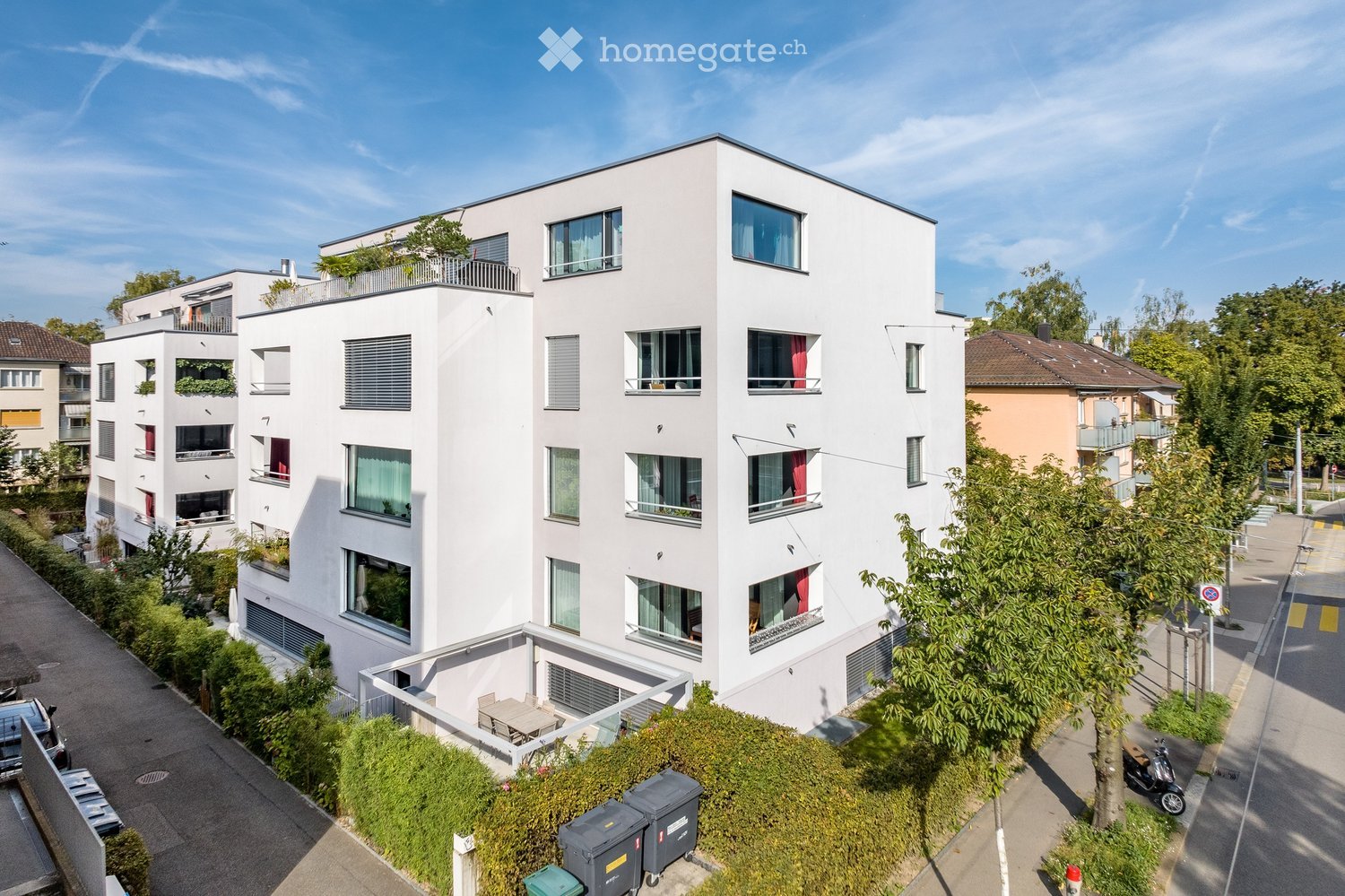 Modern white apartment building with multiple floors, balconies, and large windows. The building is surrounded by greenery and trees, with a small outdoor seating area visible on the ground floor.