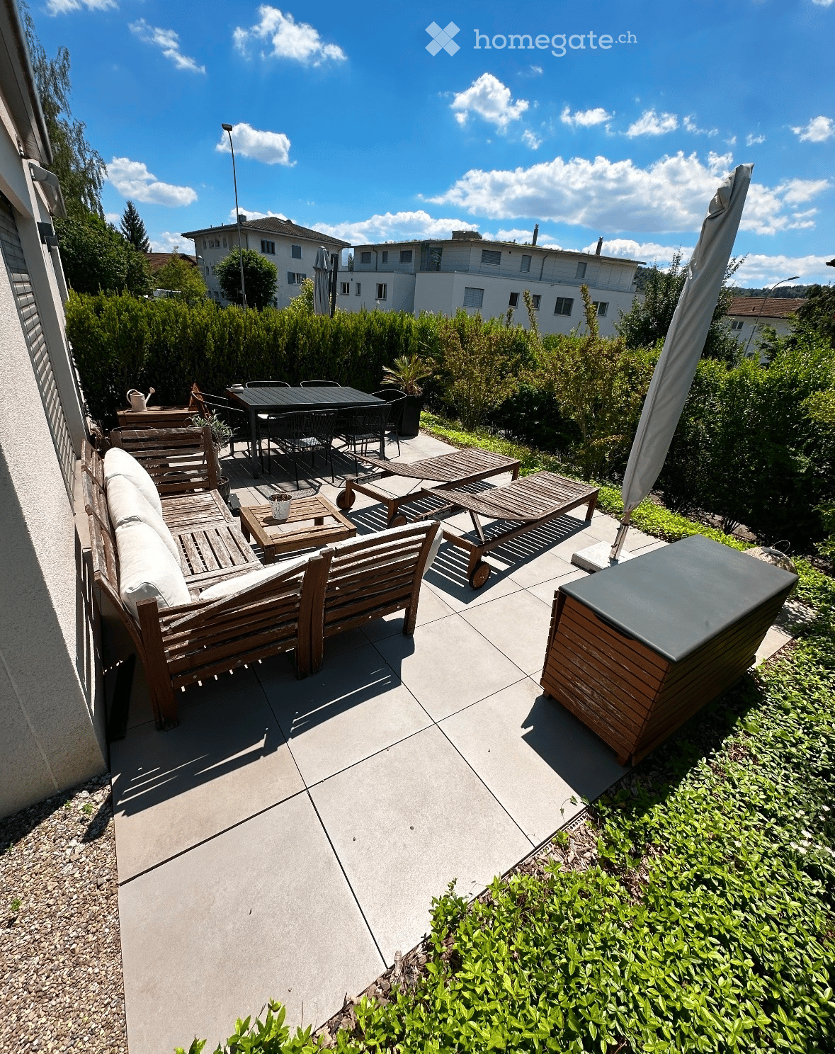 Balcony with wooden furniture including a sofa, table, chairs, and sun loungers, surrounded by greenery and a clear sky.