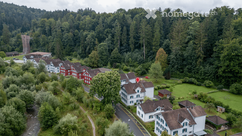 Aerial view of a residential area with multiple houses, surrounded by lush green trees and a forested backdrop. The houses are mostly two-story structures with pitched roofs, and the area includes a small parking lot and a pathway.