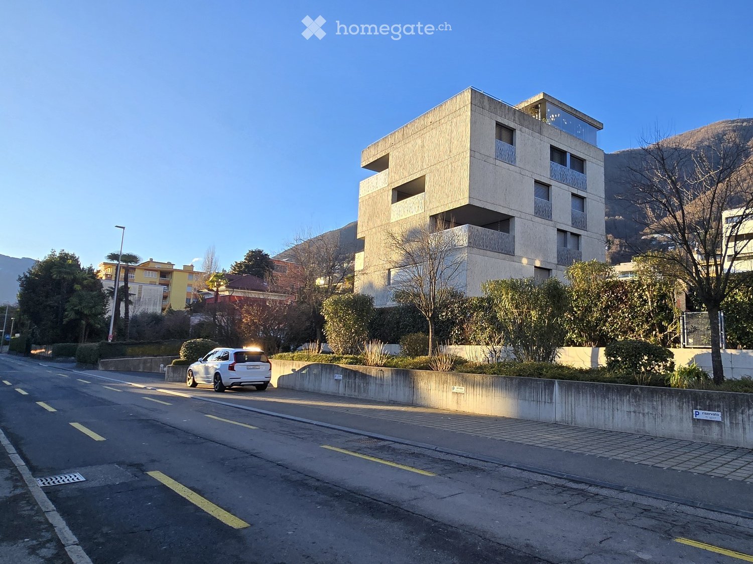 Modern building with a sloped driveway and balconies, surrounded by greenery and mountains