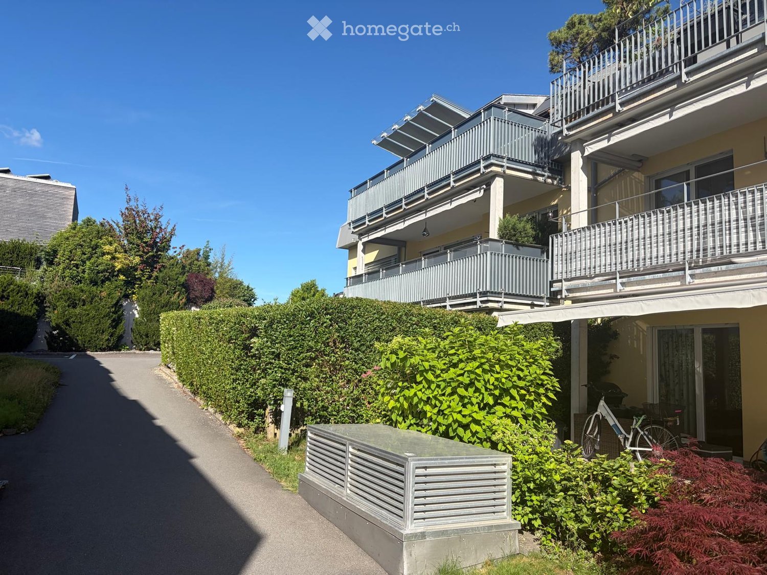 A modern multi-story residential building with balconies, surrounded by greenery and a well-maintained hedge. The building features a mix of light-colored facades and metal railings on the balconies.