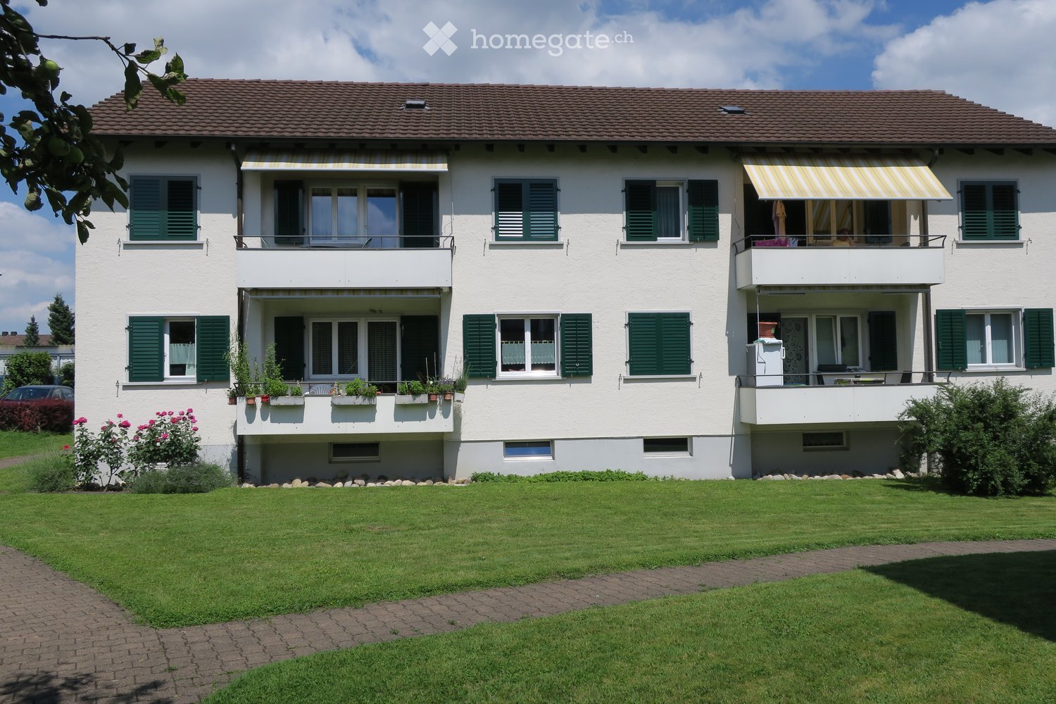2 story apartment building, white walls, brown roof, green shutters, balconies, well-maintained garden