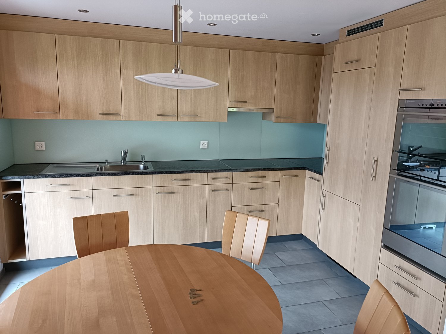 Modern kitchen with light wood cabinets, stainless steel appliances, a round wooden dining table with chairs, and a pendant light fixture.