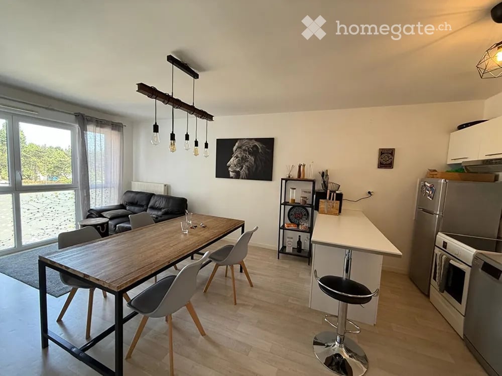 Open-plan living and dining area with a black leather sofa, wooden dining table with chairs, kitchen island with stools, and modern appliances.