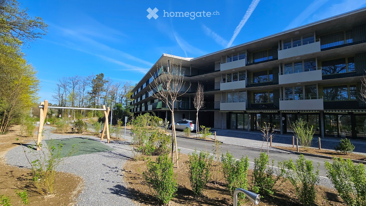 Modern multi-story residential building with balconies and large windows, surrounded by landscaped greenery and a playground area.
