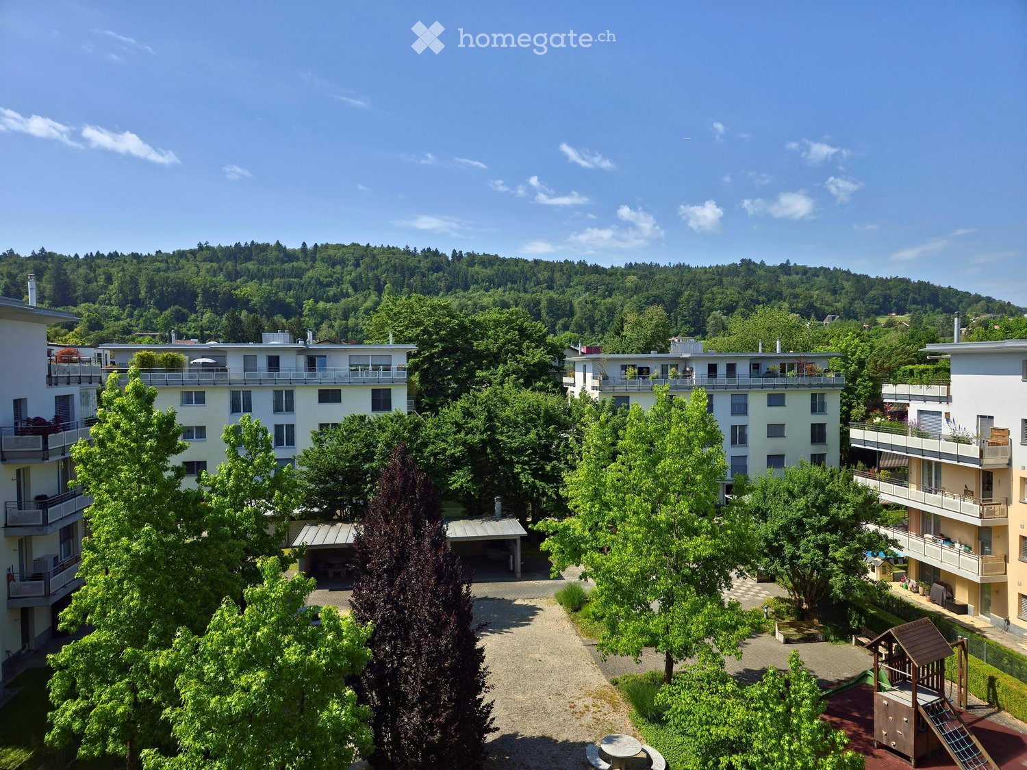 A multi-story residential building complex surrounded by lush greenery and trees, with balconies and a central courtyard featuring a playground and seating area.
