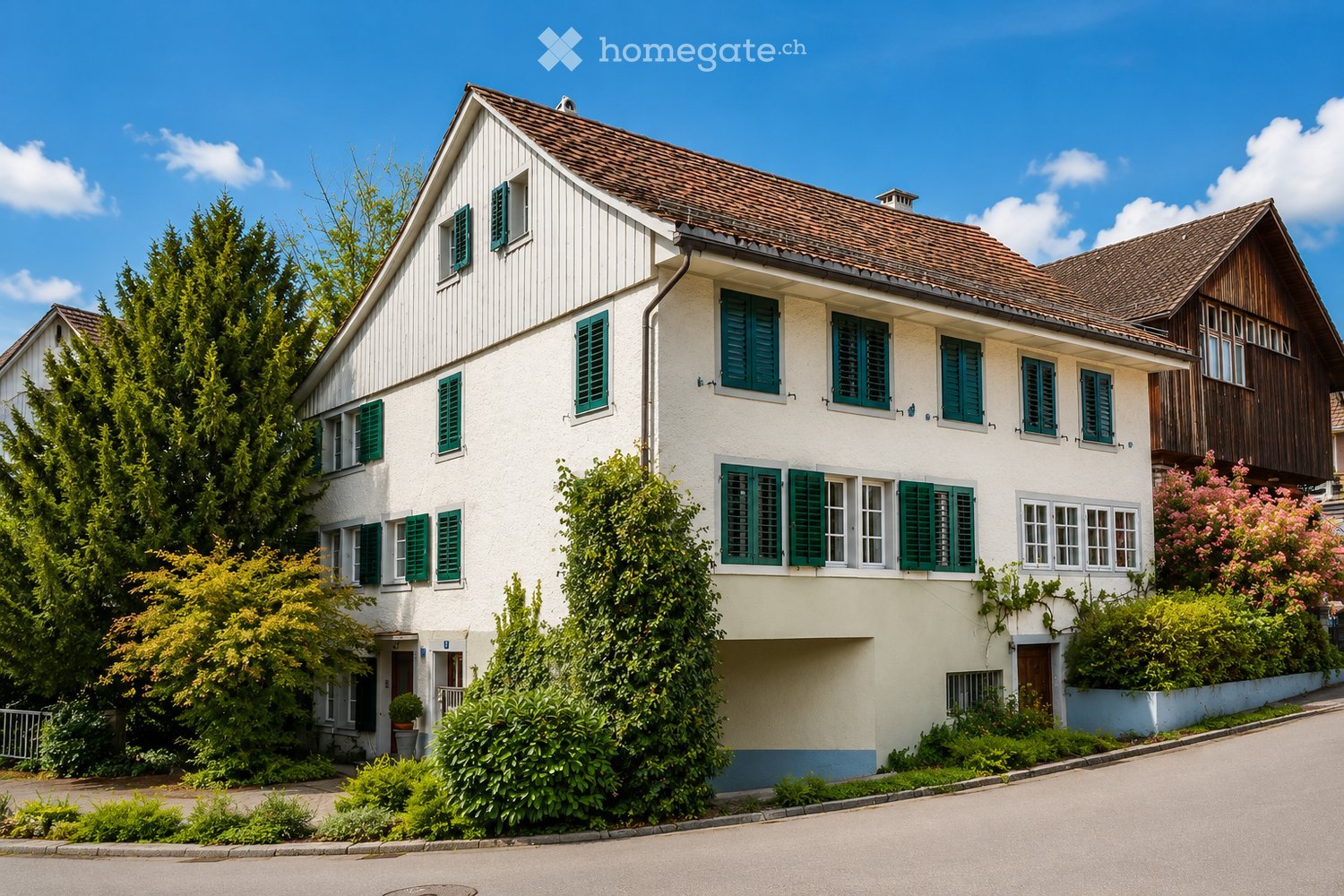 A two-story house with white walls, green shutters, and a brown tiled roof, surrounded by greenery and bushes.