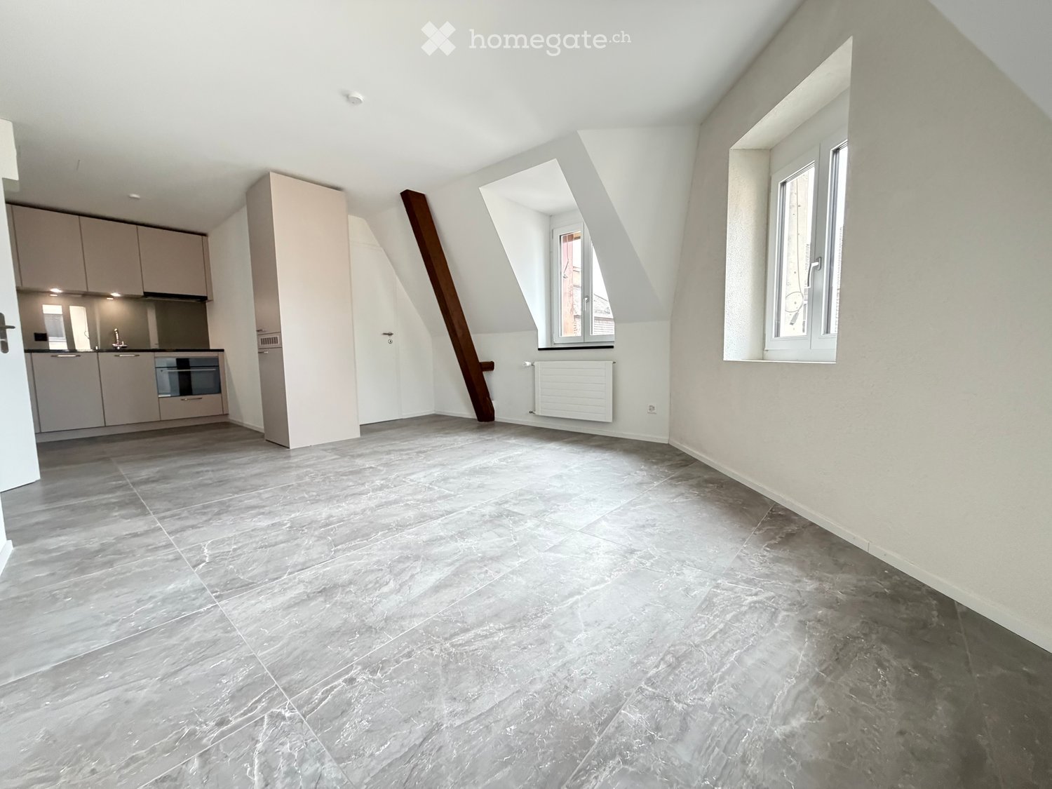 Empty room with grey tiled floor, white walls, and built-in kitchen cabinets visible in the background. The room has large windows and a slanted ceiling typical of an attic space.