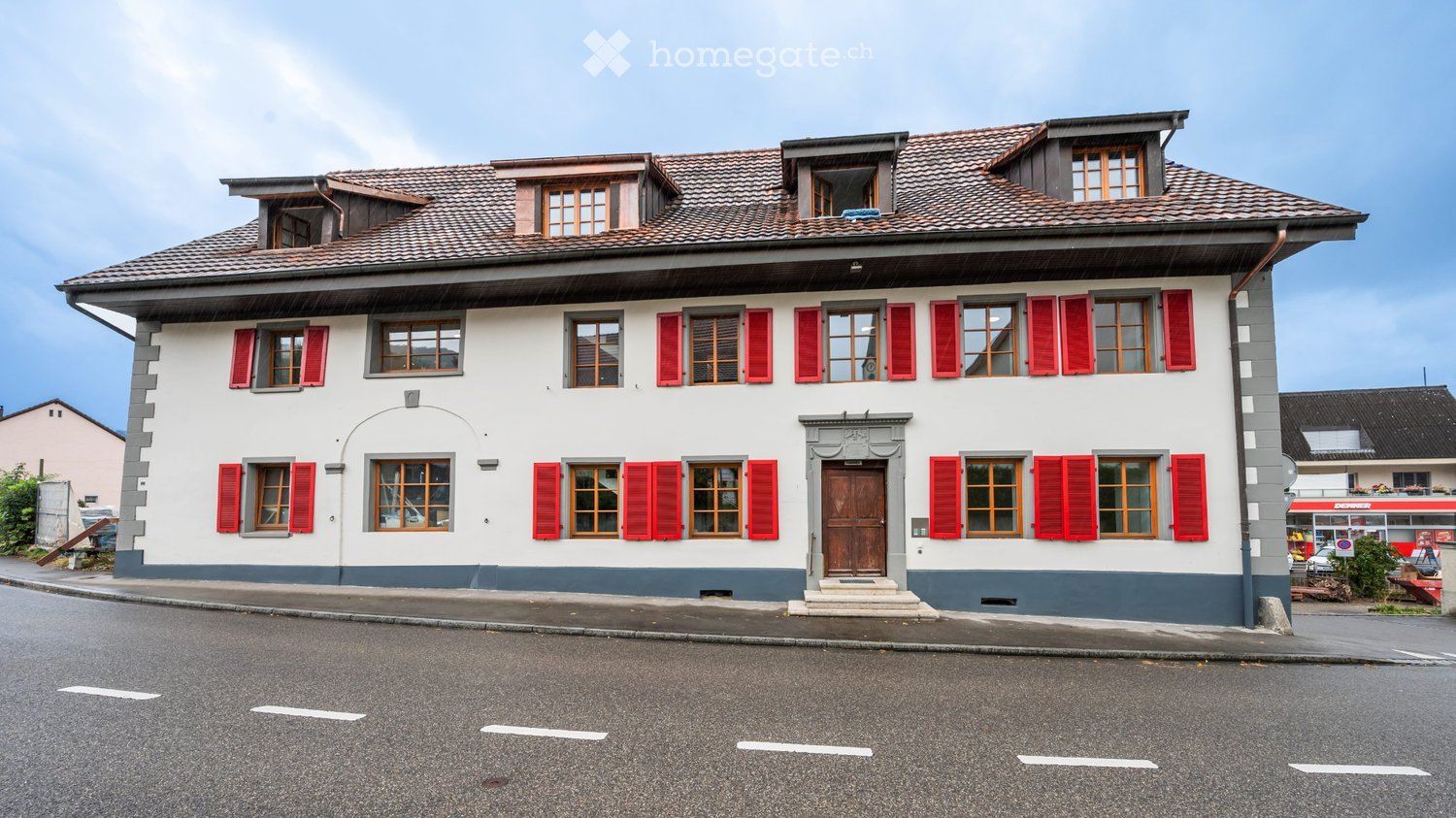Two-story house with red shutters, white walls, and a tiled roof. Features a central wooden door and multiple windows.