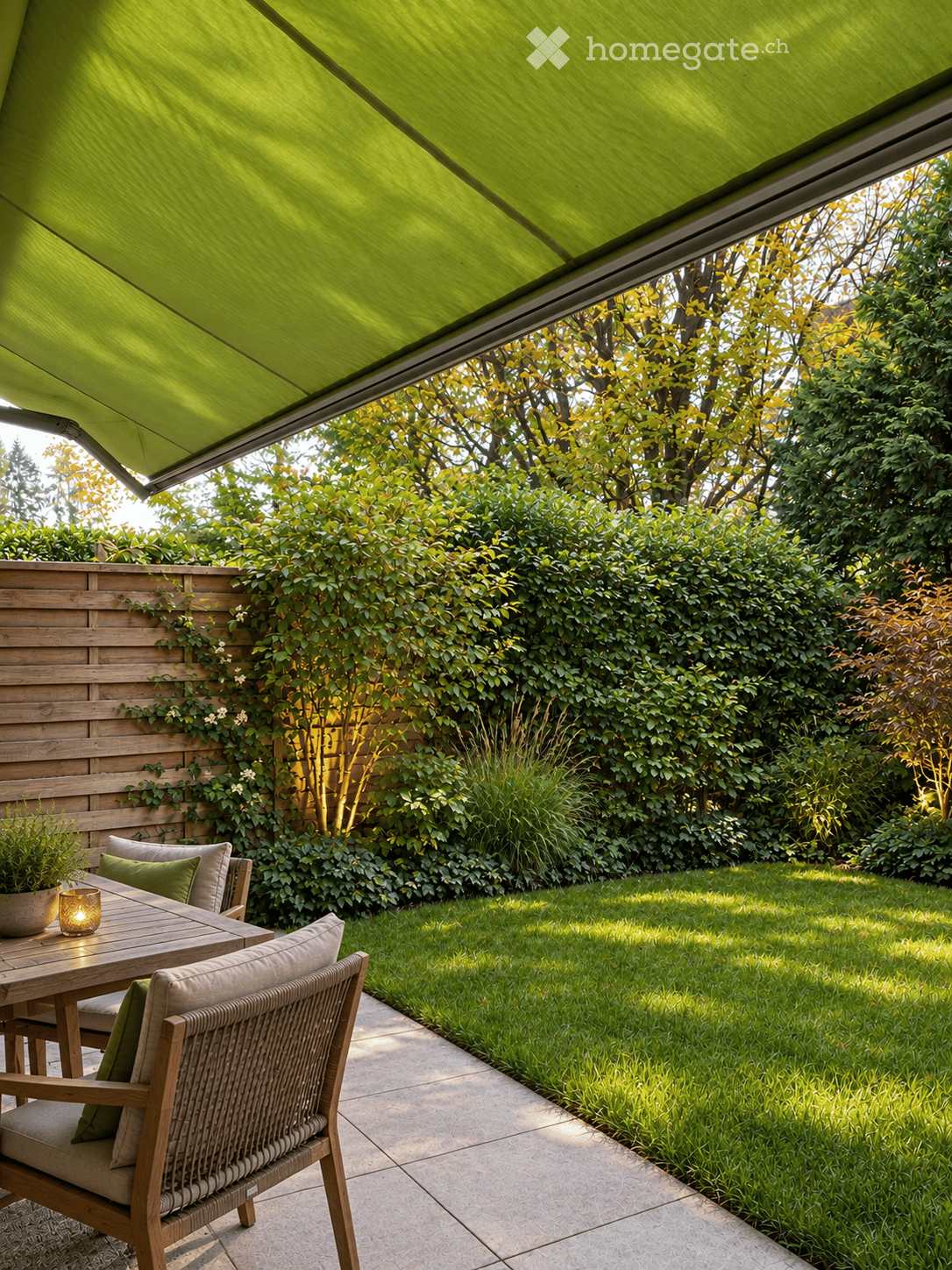 A well-maintained garden with a wooden table, chairs, and a green awning providing shade. The area is surrounded by neatly trimmed hedges and trees, with a paved pathway leading to a grassy lawn.
