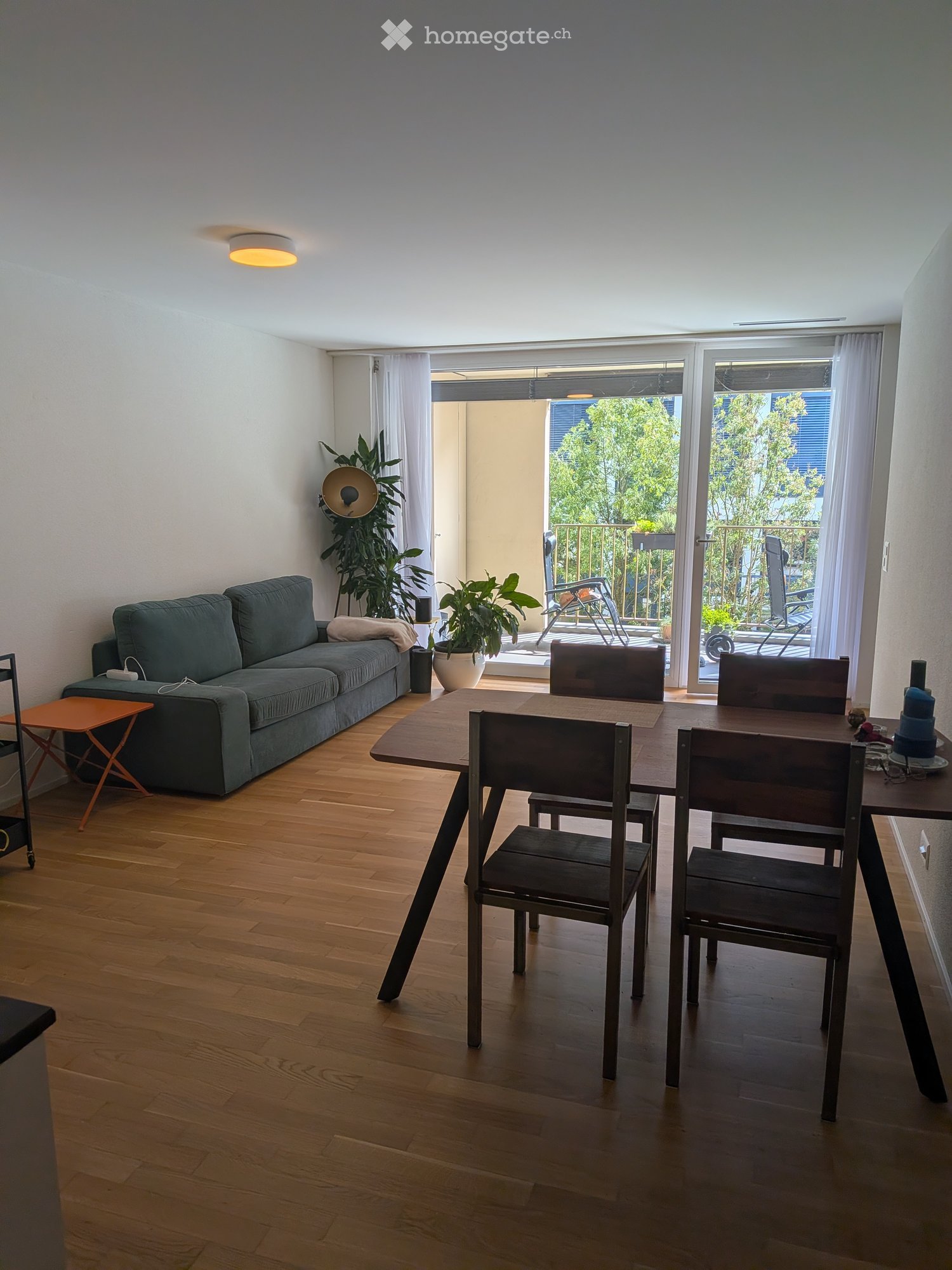Bright living and dining area with wooden flooring, a grey sofa, a small orange stool, a dining table with four chairs, and potted plants near the balcony entrance.