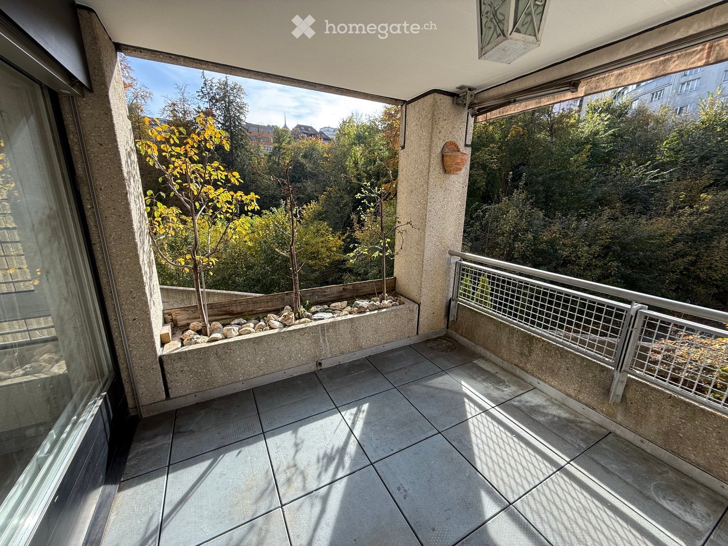 Balcony with tiled floor, metal railing, and a small garden area with rocks and plants, overlooking a green landscape.