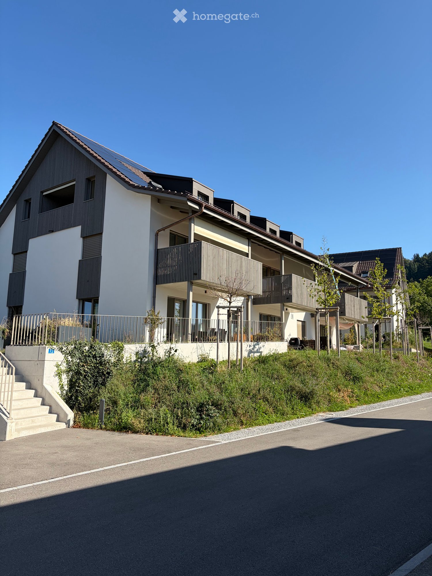 Modern two-story residential building with white and dark grey exterior, large windows, balconies, and solar panels on the roof.