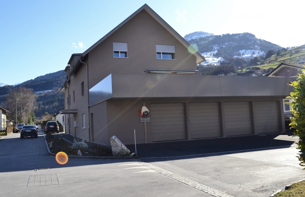 Modern two-story house with a flat roof and large garage, situated in a scenic mountainous area with snow-capped peaks in the background.