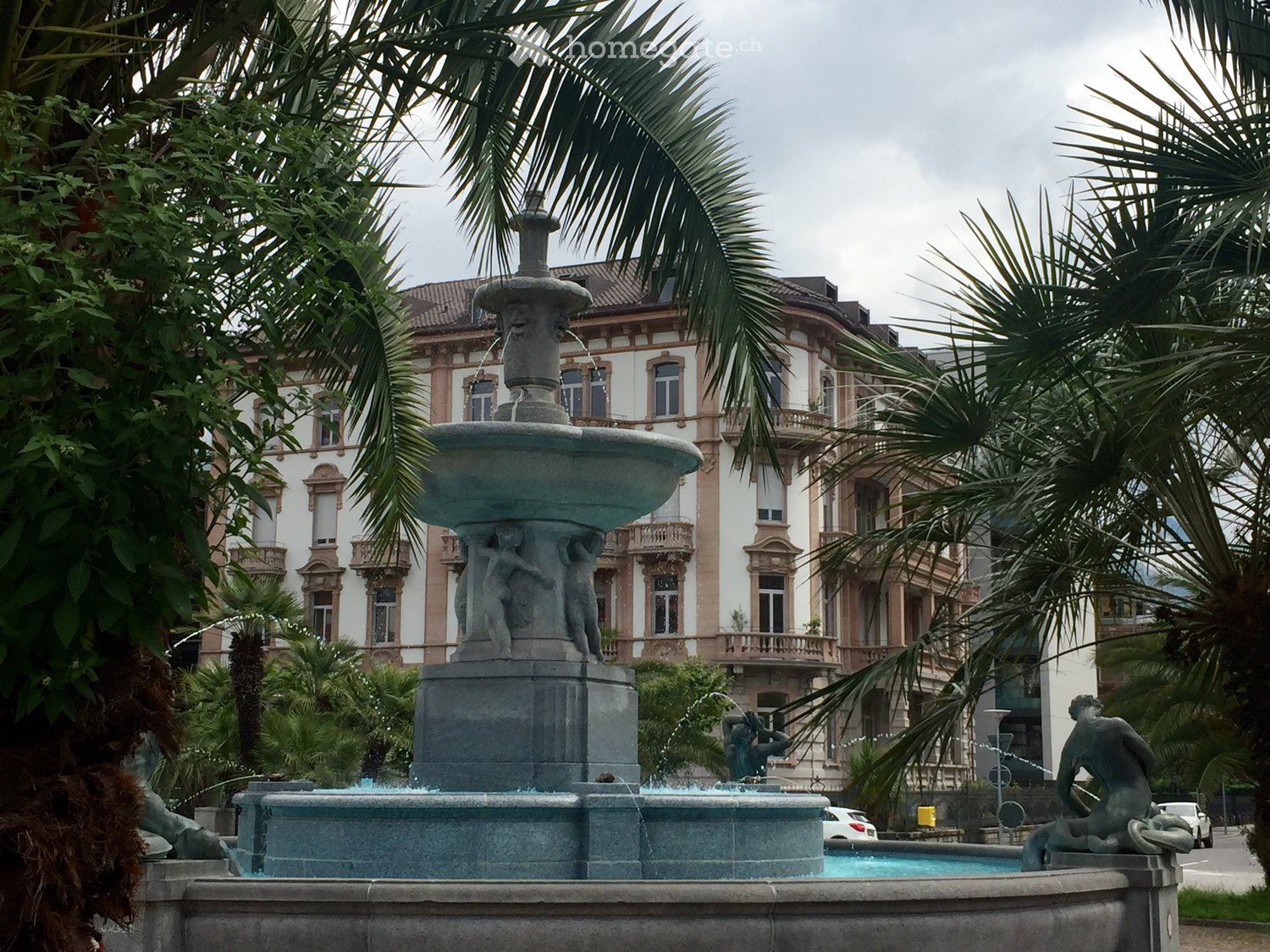 A large stone fountain with multiple tiers and sculptures, surrounded by palm trees and a grand historic building in the background.