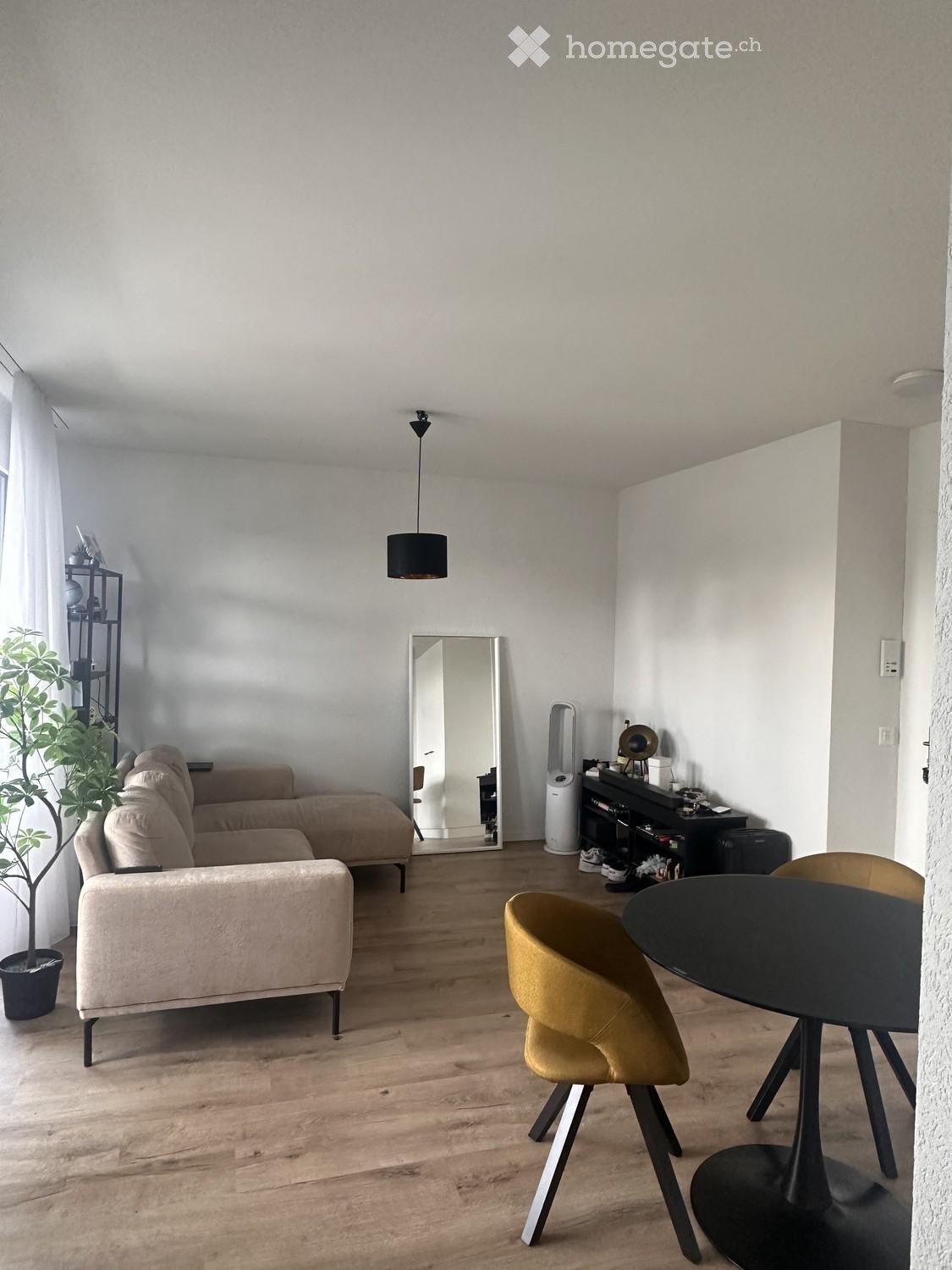 Modern living room with light wooden flooring, a beige sectional sofa, a round black dining table with yellow chairs, a large mirror, a potted plant, and a shelf with decorative items.