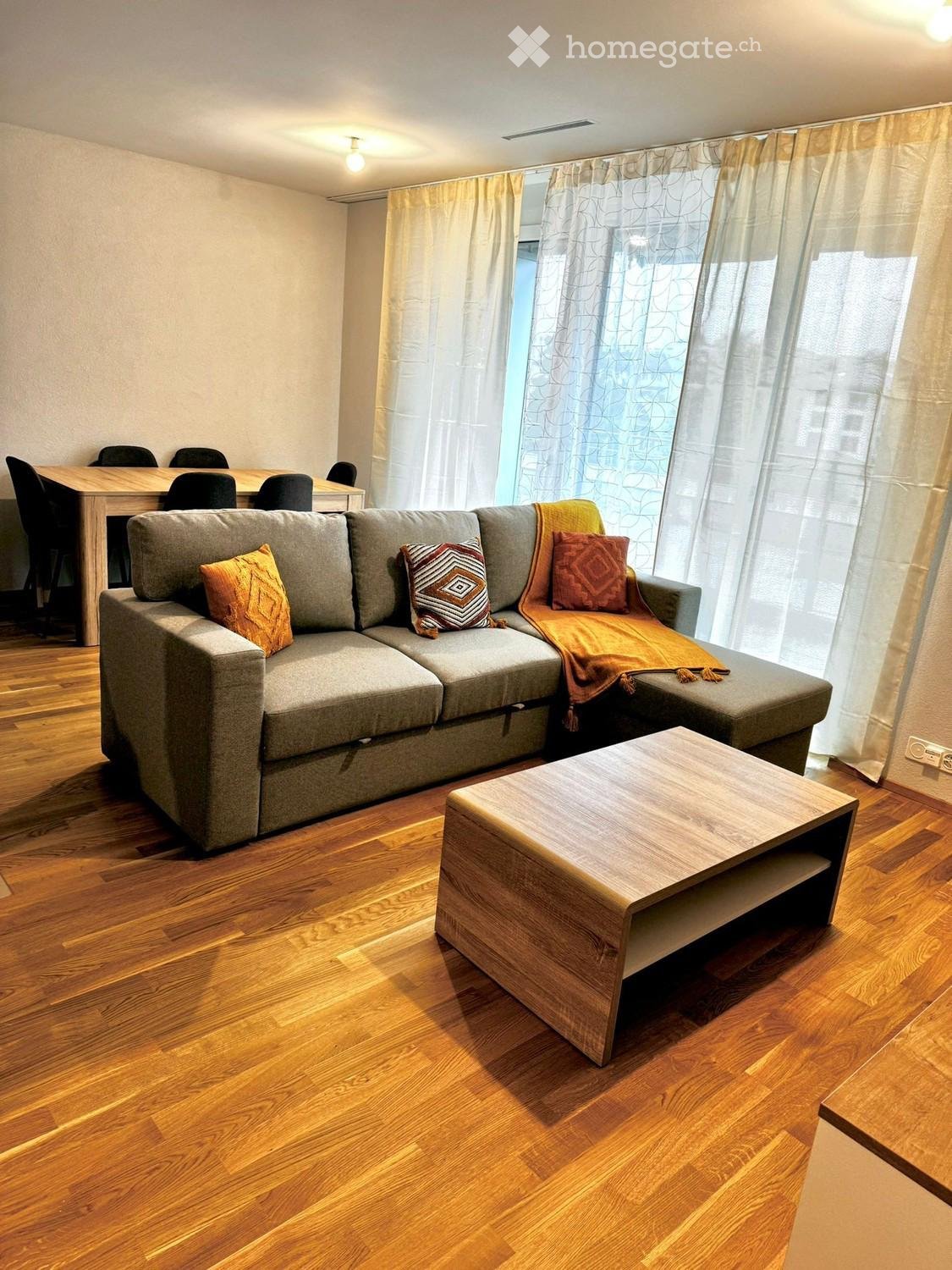 Modern living room with a grey sectional sofa, wooden coffee table, dining table with chairs, and large windows with curtains.