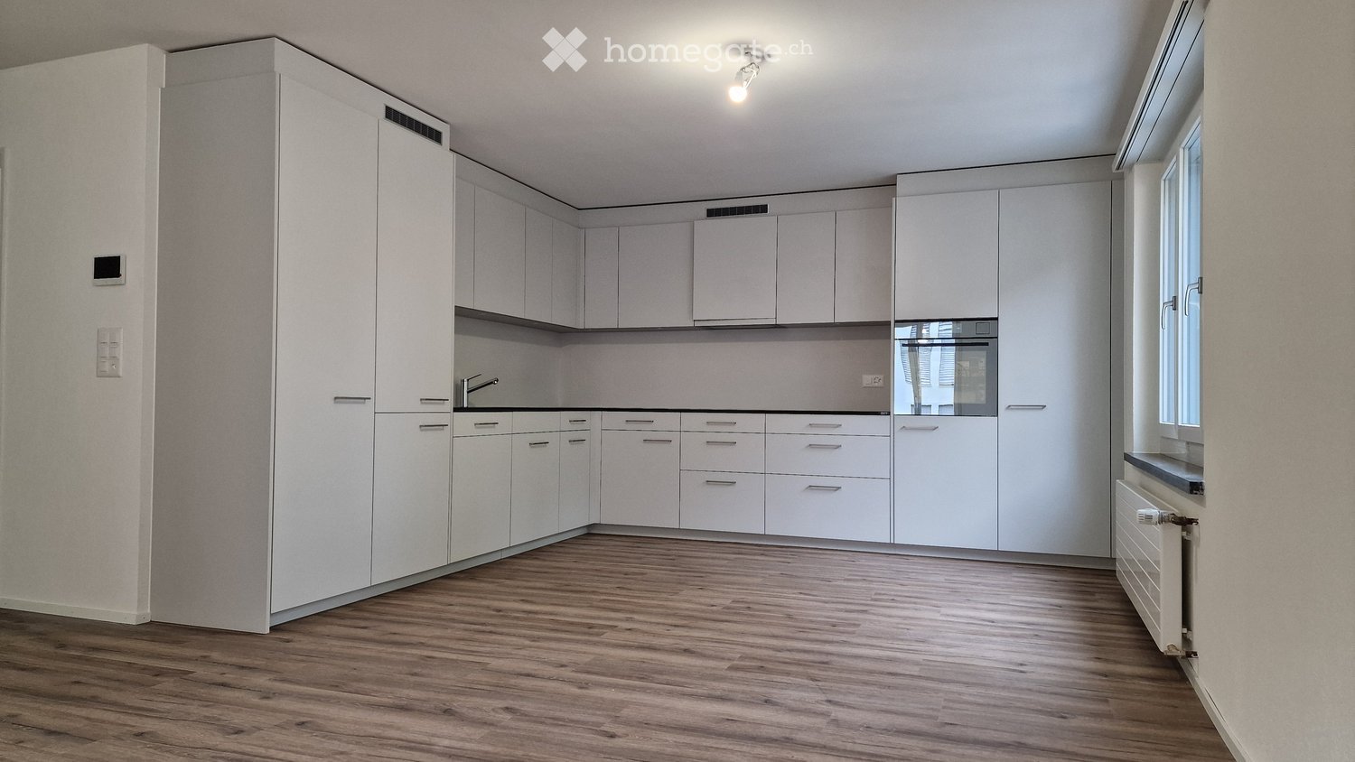 Empty kitchen with white cabinets, wooden floor, and large windows. No appliances or furniture are present.