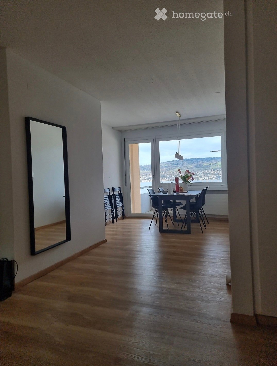 Bright, open dining area with wooden flooring, large windows offering a scenic view, a dining table with chairs, and a decorative flower vase on the table.