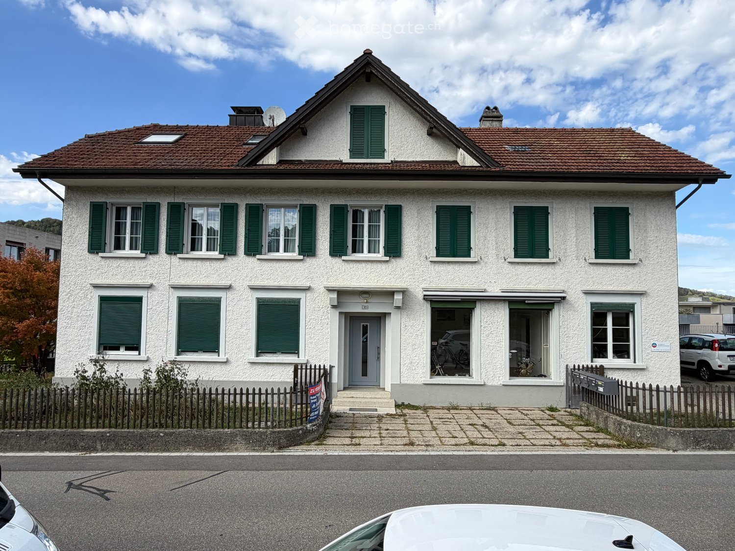 Two-story house with white exterior, green shutters, red-tiled roof, and a small balcony area visible through the windows.