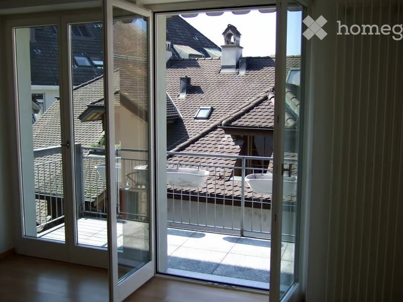Balcony with metal railing, overlooking neighboring rooftops and chimneys. The balcony is accessible through large glass doors from the interior.
