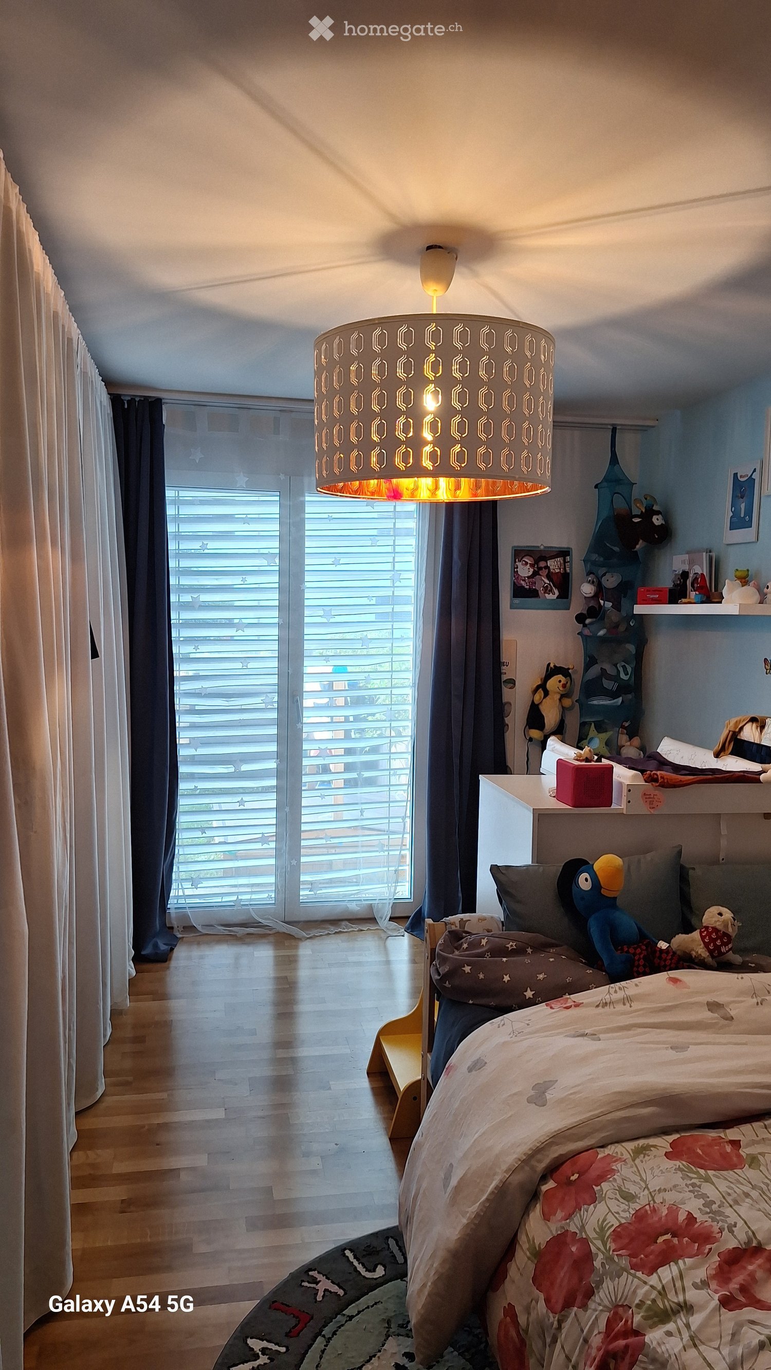 Brightly lit bedroom with wooden flooring, large window with blinds, bed with floral bedspread, decorative pillows, stuffed toys, and a hanging lamp with a geometric shade.