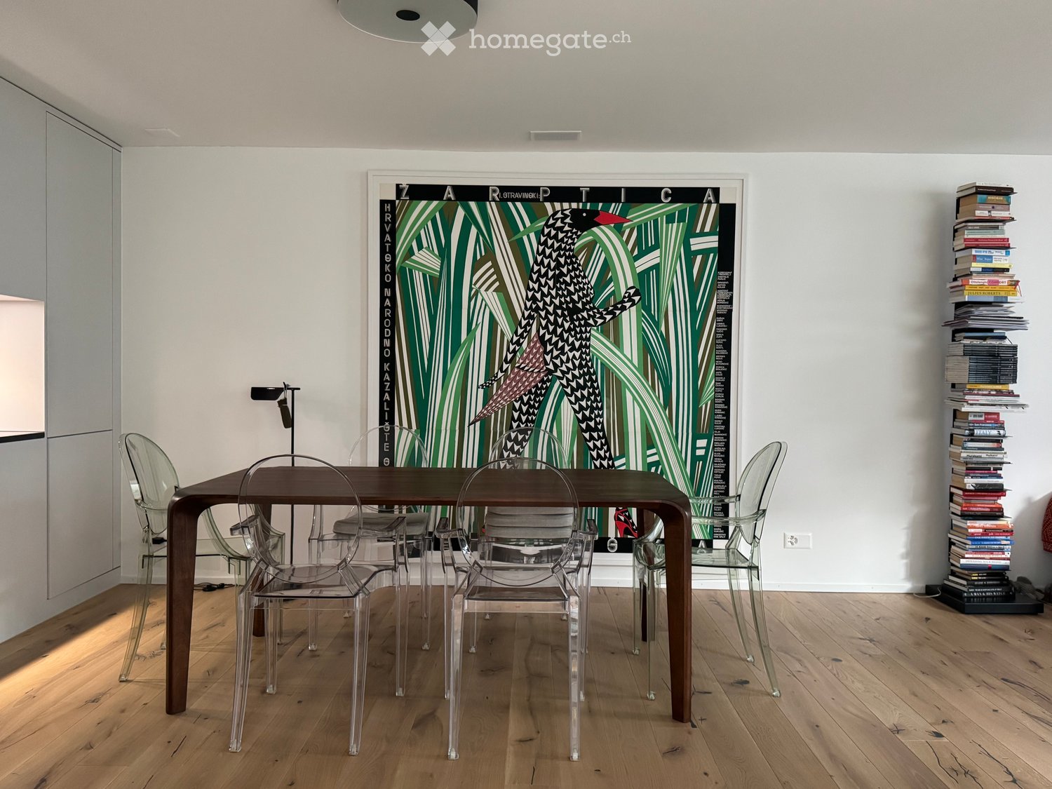 Dining area with a wooden table, clear chairs, and a large wall-mounted artwork. Books are stacked on the right side.