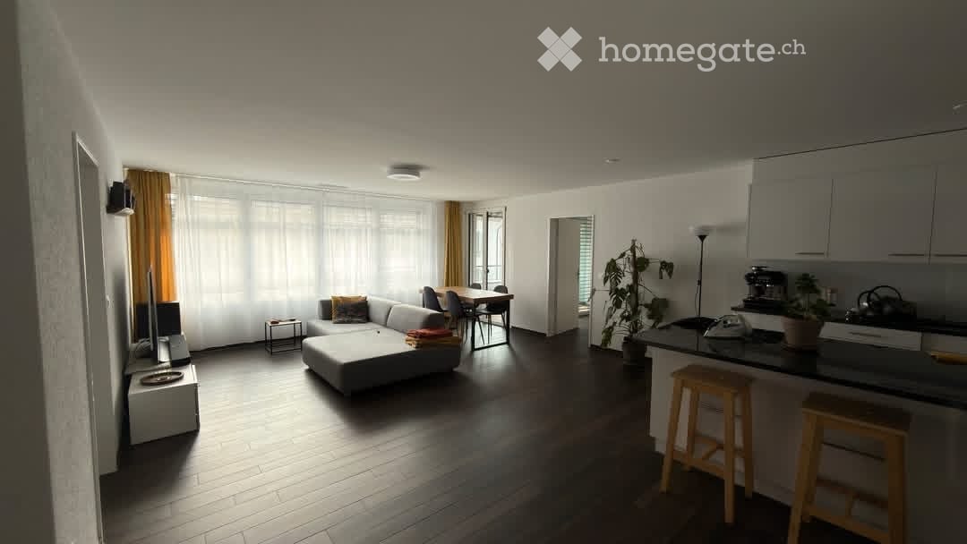 Open-plan living and dining area with a grey sectional sofa, coffee table, dining table with chairs, kitchen island with stools, and a potted plant. The space features large windows with sheer curtains and wooden flooring.