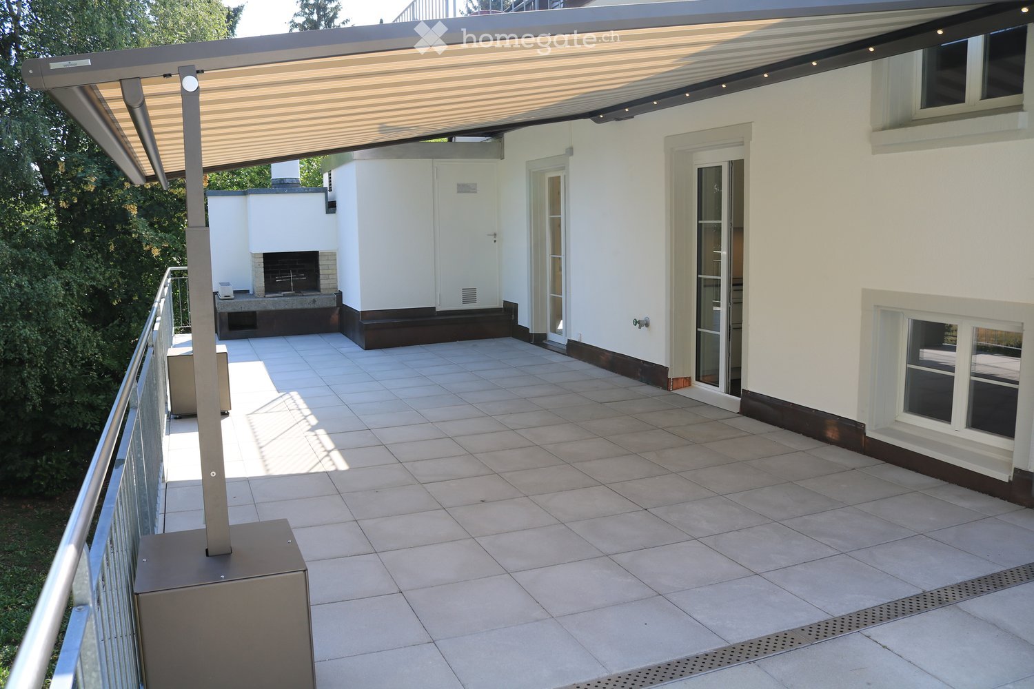 Balcony with tiled floor, metal railing, covered patio area with a retractable awning, and a fireplace in the background.