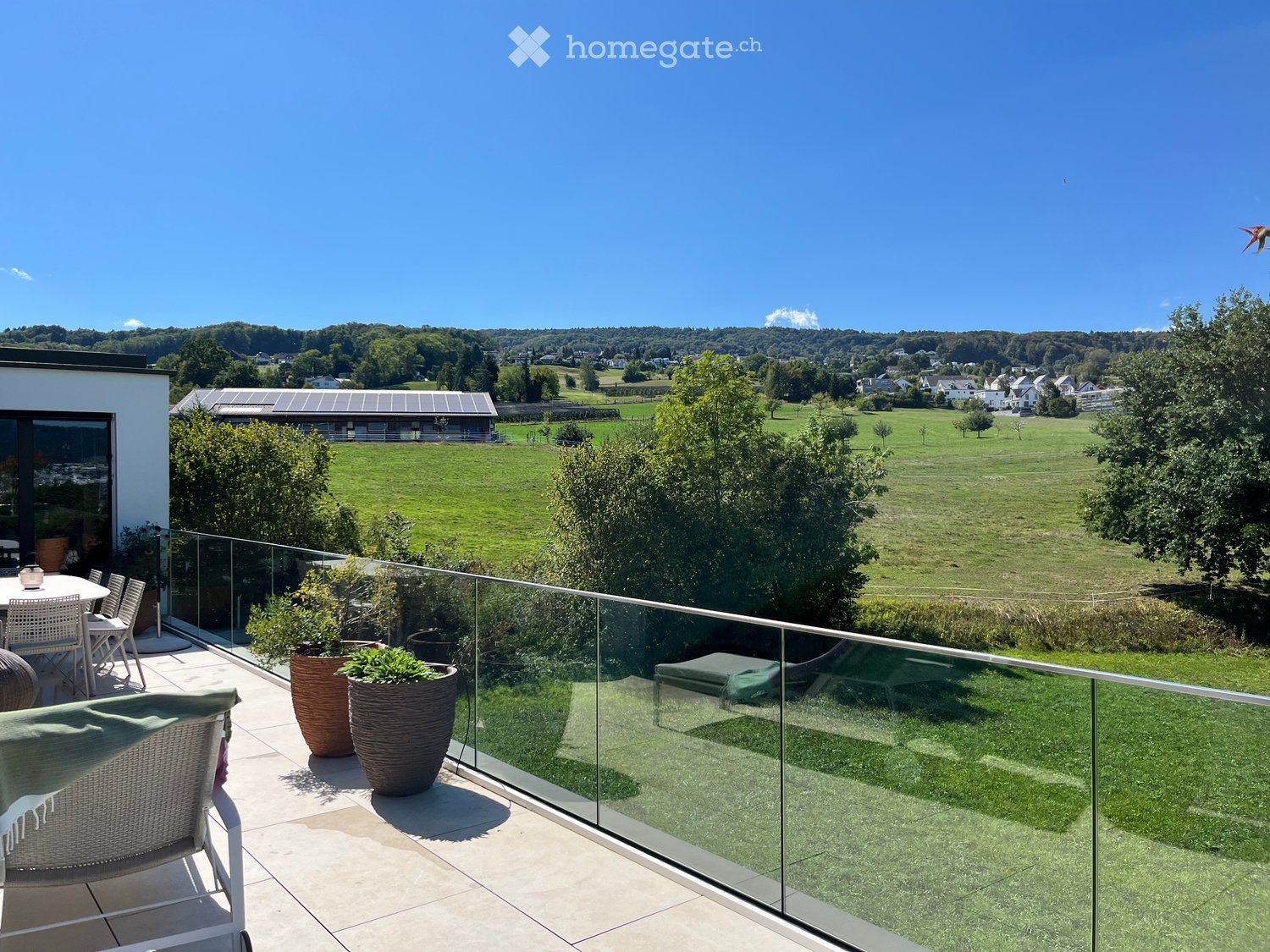 Balcony with glass railing, overlooking a green field and distant houses, furnished with a chair, table, and potted plants.