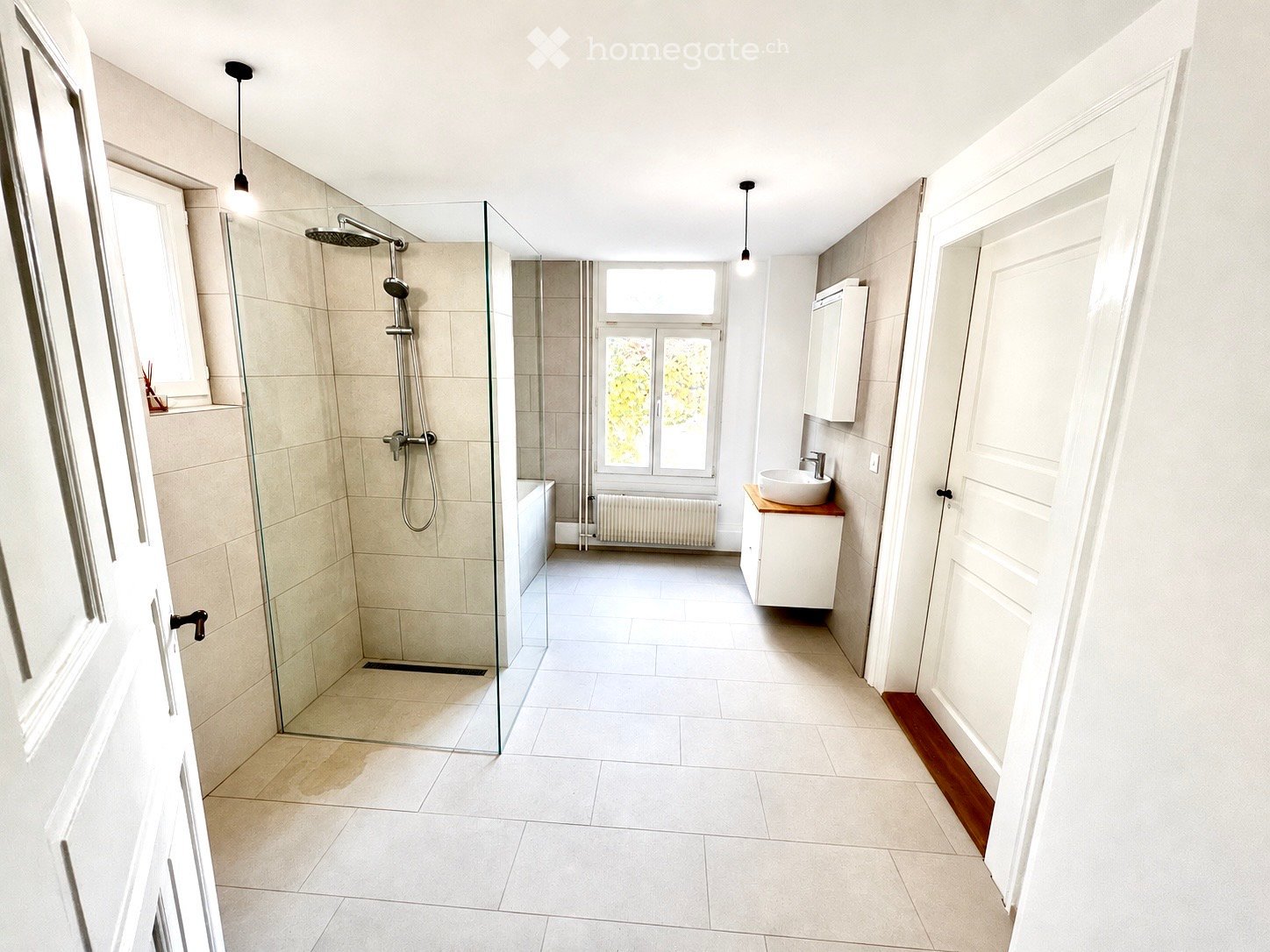Modern bathroom with a glass-enclosed shower, a white sink on a wooden vanity, and a white cabinet above it. The space features light-colored tiles and a large window providing natural light.