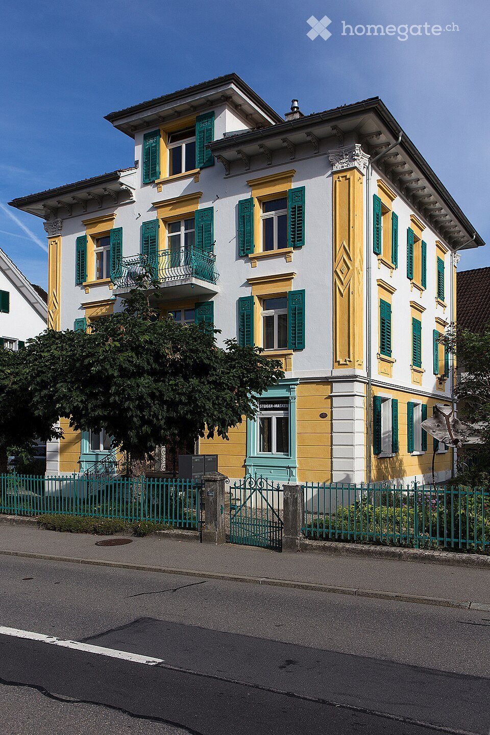 A multi-story residential building with white and yellow facade, green shutters, and a green metal fence in front.