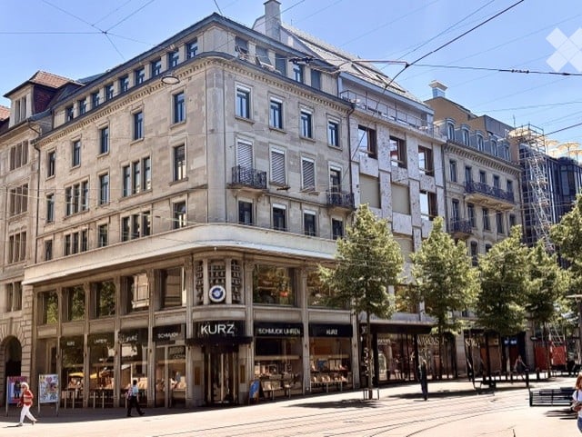 A multi-story urban building with classic architecture, large windows, and a storefront labeled "KURZ" at the ground level. The building features ornate detailing and is situated on a busy street with pedestrians and trees.