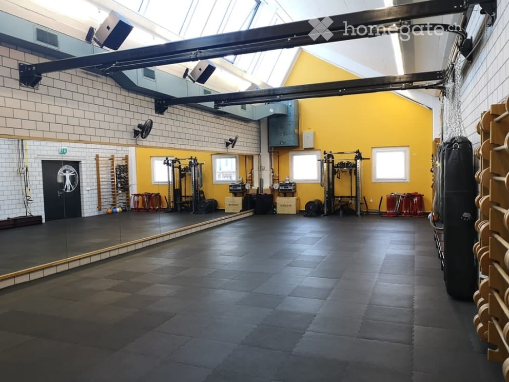 A spacious gym room with a dark rubber floor, large mirror wall, punching bag, weight equipment, and storage racks. The room features yellow accent walls and ample natural light from skylights and windows.