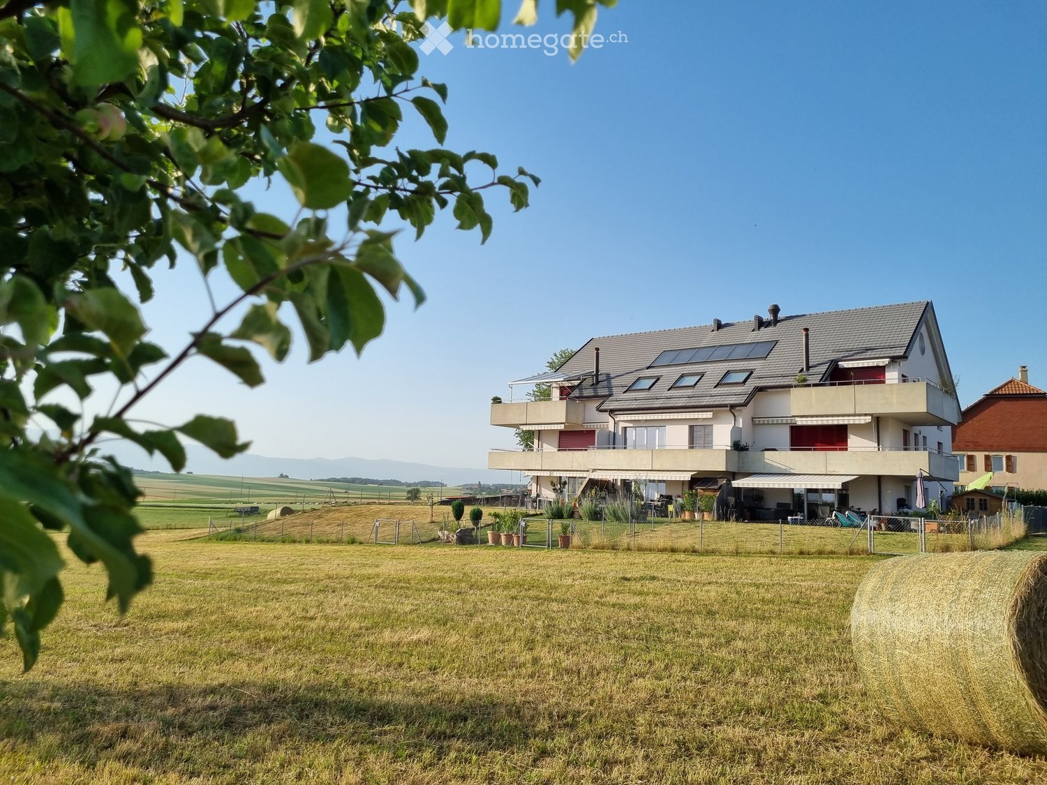 Modern two-story house with a grey roof, white walls, and solar panels, situated in a rural area with a spacious lawn and surrounding fields.
