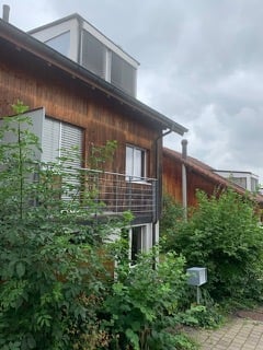 A two-story wooden house with a modern design, featuring large windows, a balcony, and a metal railing. The building is surrounded by greenery and has a paved pathway leading to the entrance.