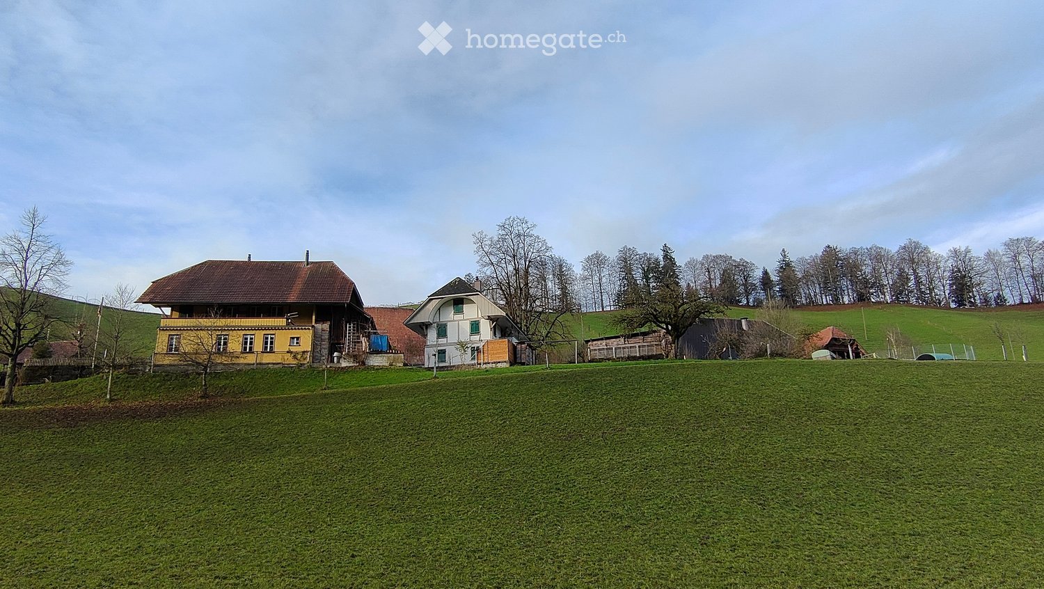 A two-story house with a yellow exterior and a brown roof, situated on a grassy field with trees and hills in the background.