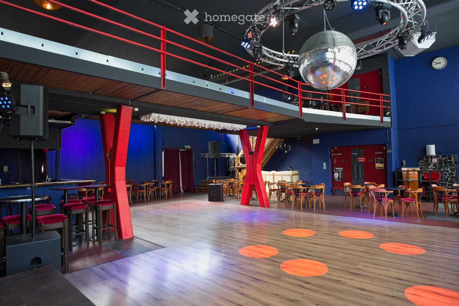 A spacious, modern dance studio or event space with wooden flooring, red accent pillars, and a balcony level. The main area features multiple tables and chairs arranged for seating, with a large disco ball hanging from the ceiling and colorful spotlights.