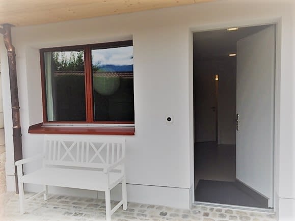 White wooden bench placed near a window with a view of greenery. The entrance door is open, revealing a dimly lit interior space.