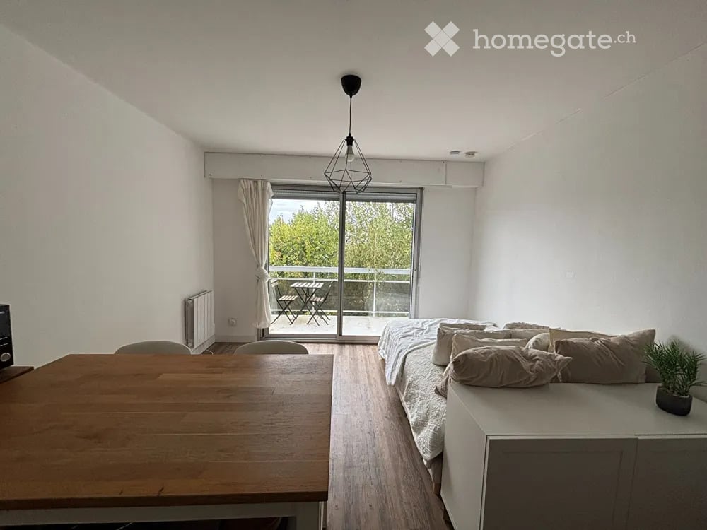A modern, well-lit living space with a sofa, a bed, a dining table, and chairs. The room features large windows with a view of a balcony area with outdoor furniture.