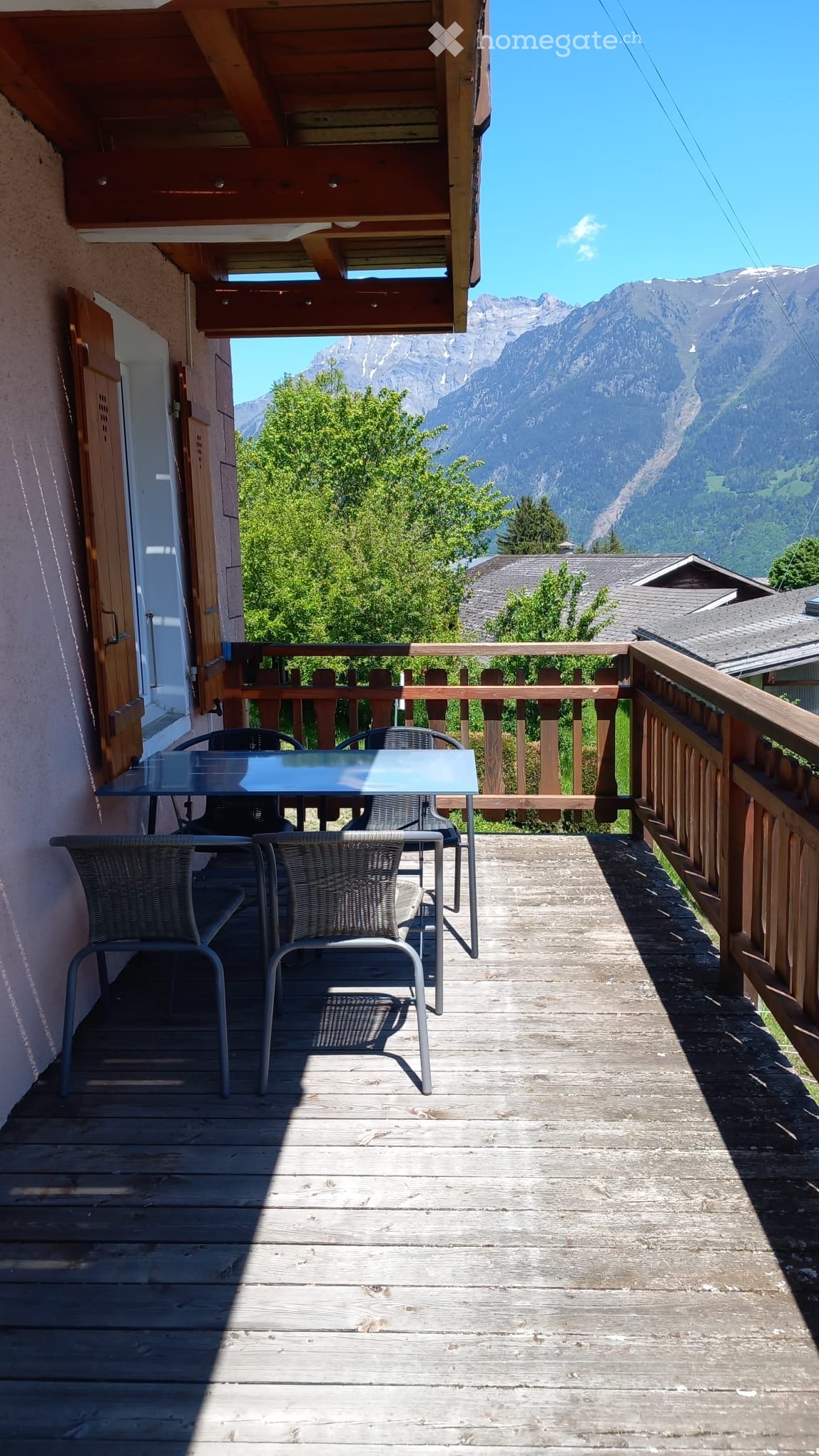 Balcony with wooden decking, glass table, and wicker chairs, overlooking a mountain view.