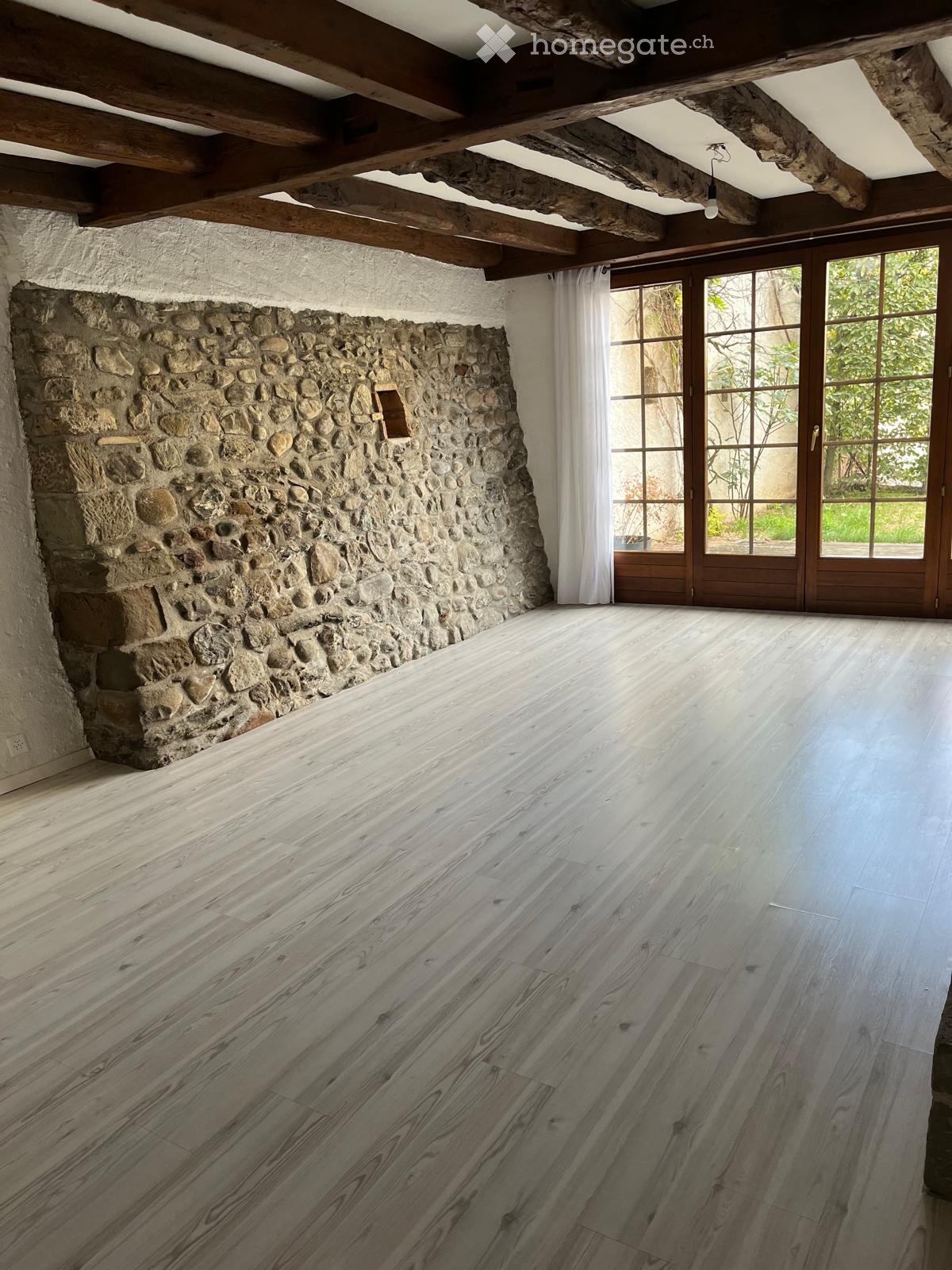 Empty room with exposed wooden beams, stone wall, large windows with white curtains, and polished wooden floor.