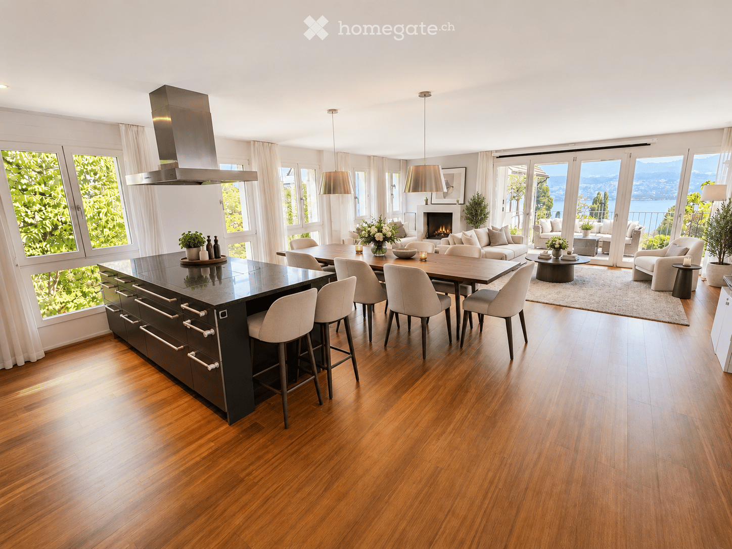 Open-plan kitchen and living area with hardwood flooring, large windows, a central island, dining area, and a living area with a sofa and fireplace. The space features modern lighting and ample natural light.