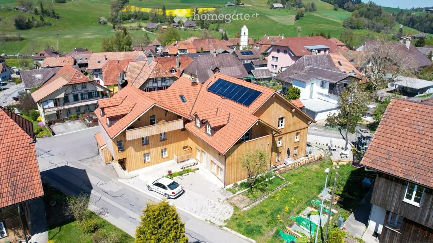Aerial view of a residential area with multiple houses, featuring a prominent wooden house with solar panels on the roof, a driveway, and a parked car.