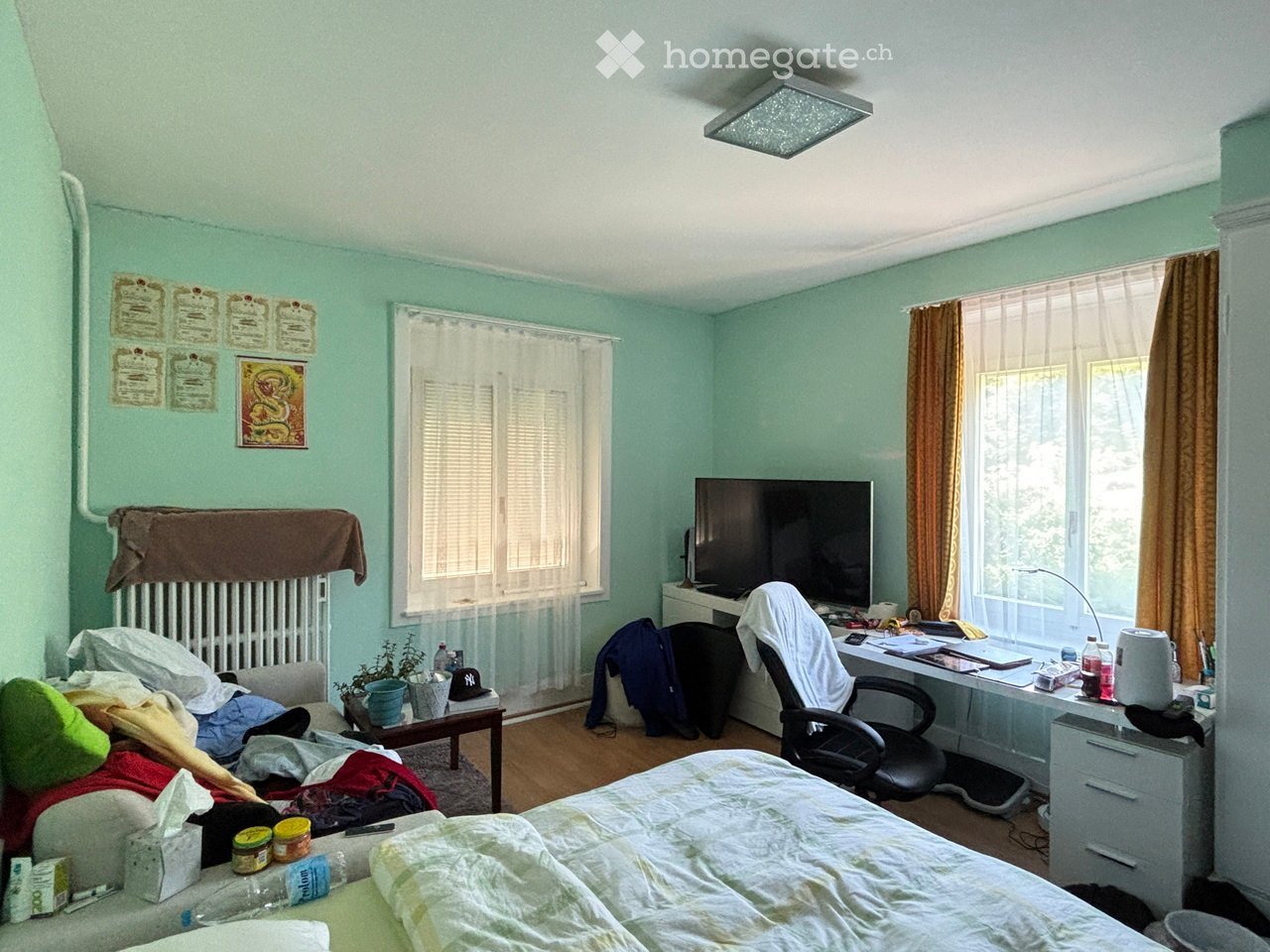 A bedroom with a bed, desk, chair, screen, small table, and various personal items. The room features light green walls, wooden flooring, and large windows with curtains.